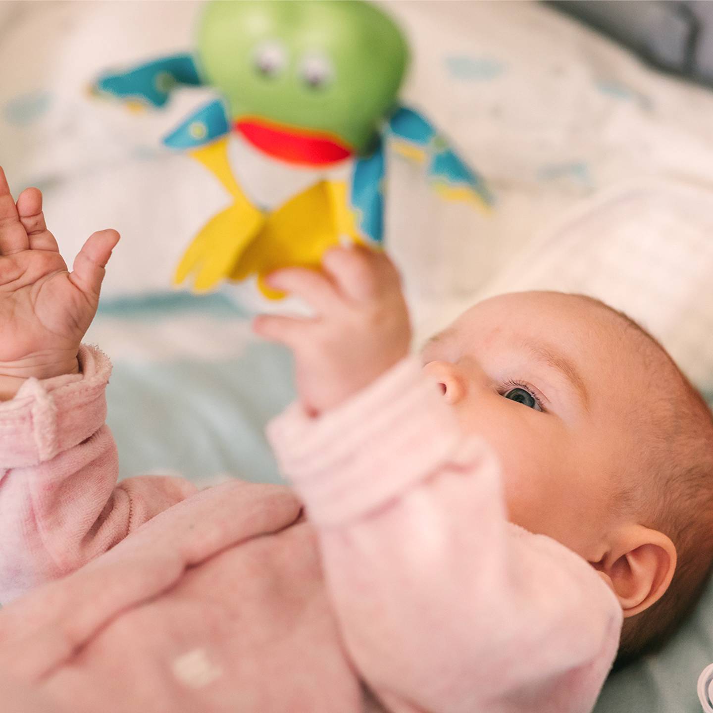 Baby im rosa Strampler greift nach einem bunten Plüschfrosch, während es auf einer weichen Oberfläche liegt.