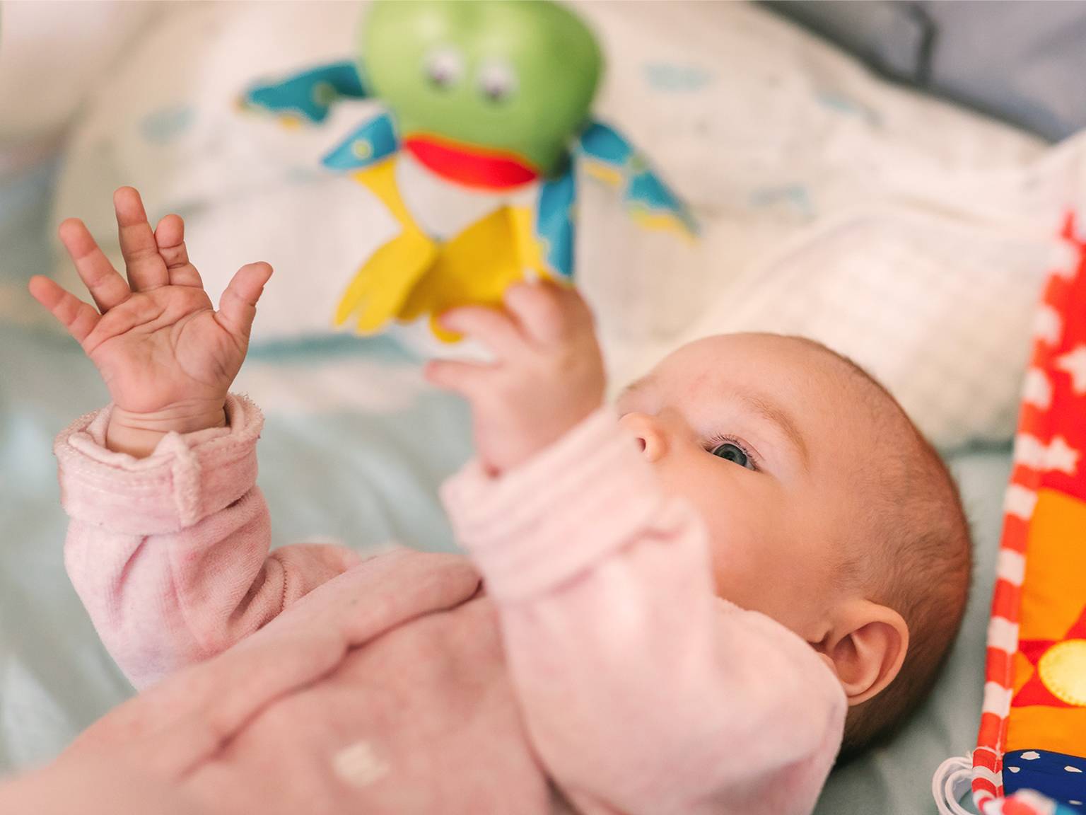 Baby im rosa Strampler greift nach einem bunten Plüschfrosch, während es auf einer weichen Oberfläche liegt.