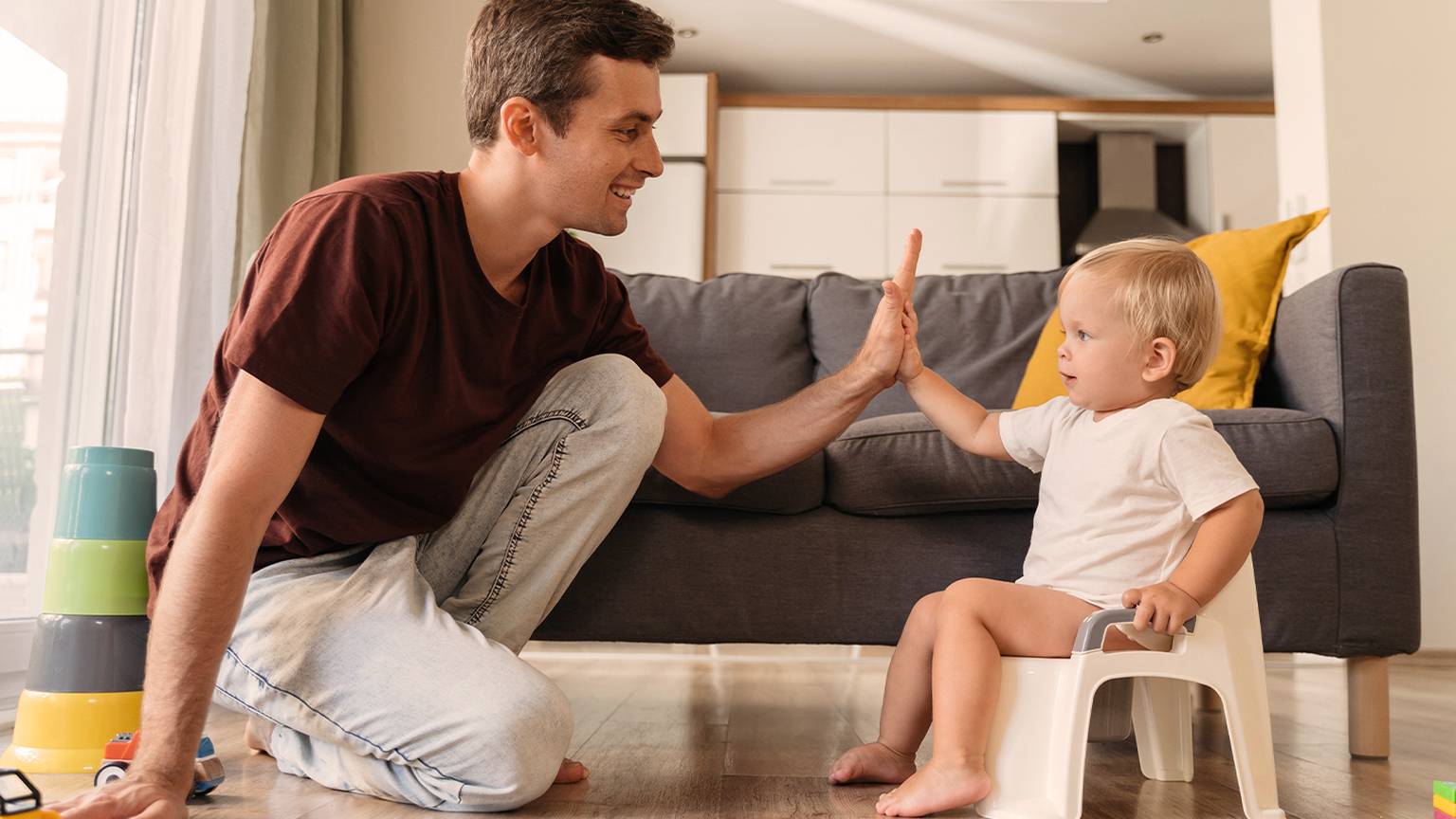 Vater und Kleinkind lächeln und geben sich in einem Wohnzimmer ein High-Five, im Hintergrund sind Spielzeuge und ein Sofa zu sehen.