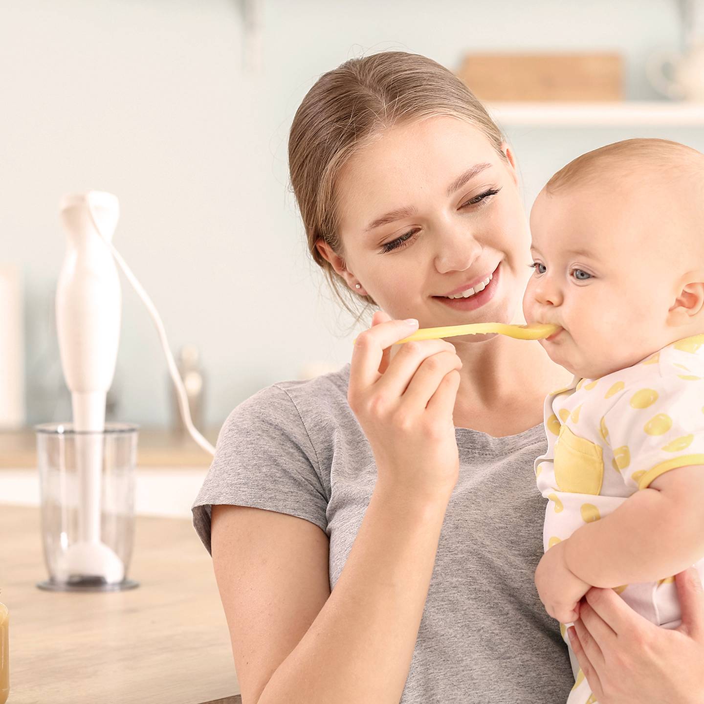 Eine lächelnde Frau füttert ein Baby in der Küche mit einem Löffel. Das Baby trägt ein gepunktetes Outfit. Auf der Arbeitsfläche stehen ein Mixer und Äpfel.