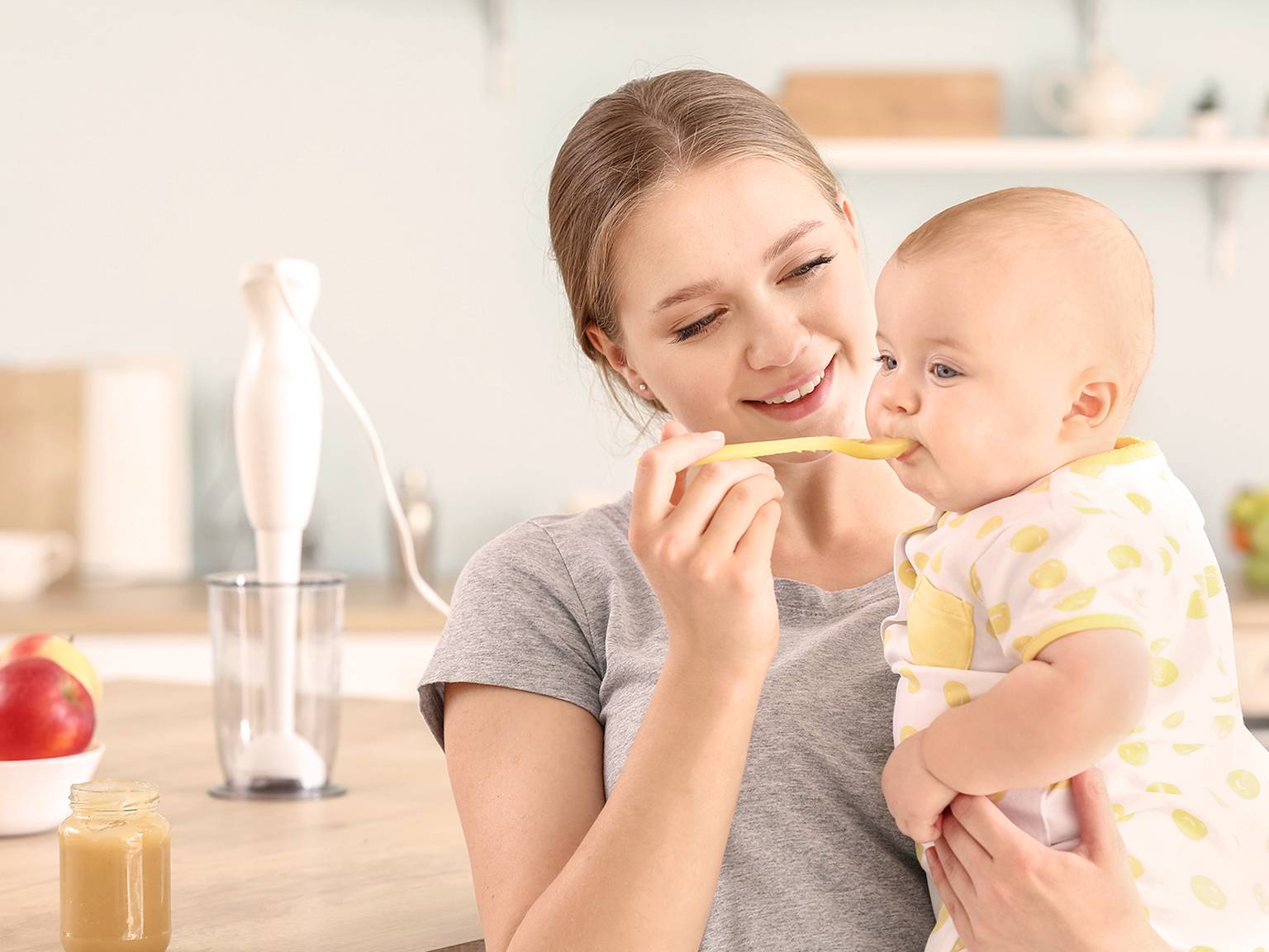 Eine lächelnde Frau füttert ein Baby in der Küche mit einem Löffel. Das Baby trägt ein gepunktetes Outfit. Auf der Arbeitsfläche stehen ein Mixer und Äpfel.