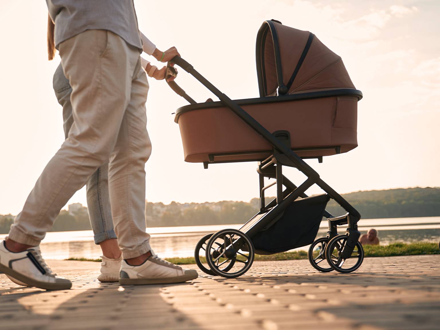 Ein Paar geht mit einen braunen Kinderwagen am Seeufer entlang. Im Hintergrund geht die Sonne unter und wirft einen warmen Schein auf die Landschaft.