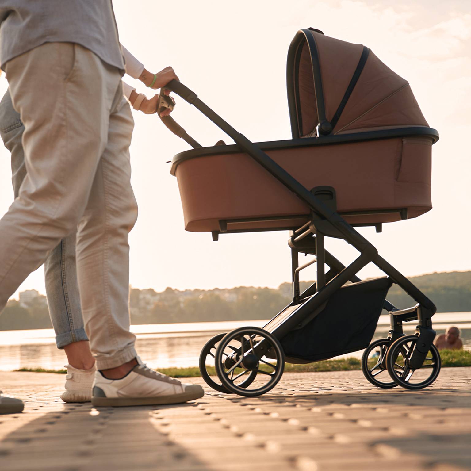 Ein Paar geht mit einen braunen Kinderwagen am Seeufer entlang. Im Hintergrund geht die Sonne unter und wirft einen warmen Schein auf die Landschaft.