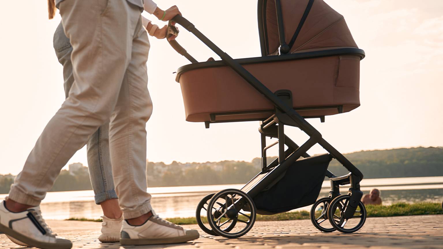 Ein Paar geht mit einen braunen Kinderwagen am Seeufer entlang. Im Hintergrund geht die Sonne unter und wirft einen warmen Schein auf die Landschaft.