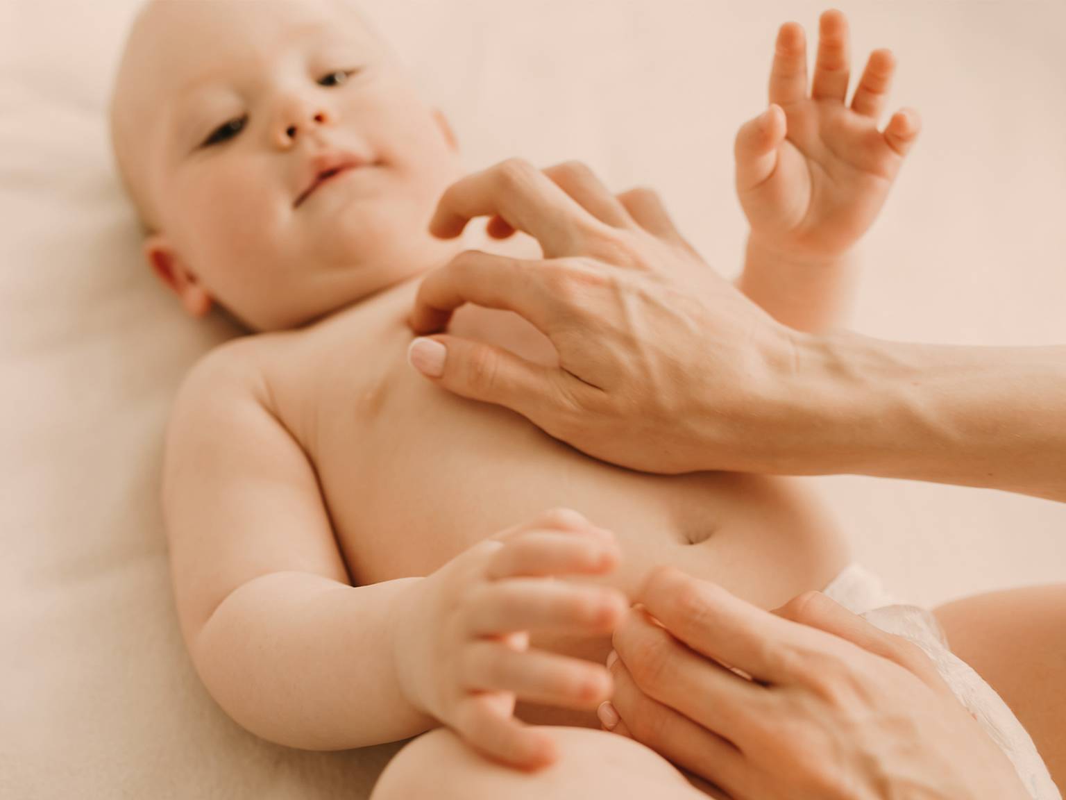 Ein Baby liegt mit freudigem Gesichtsausdruck auf einer weichen Oberfläche und wird sanft von den Händen eines Erwachsenen gekitzelt.