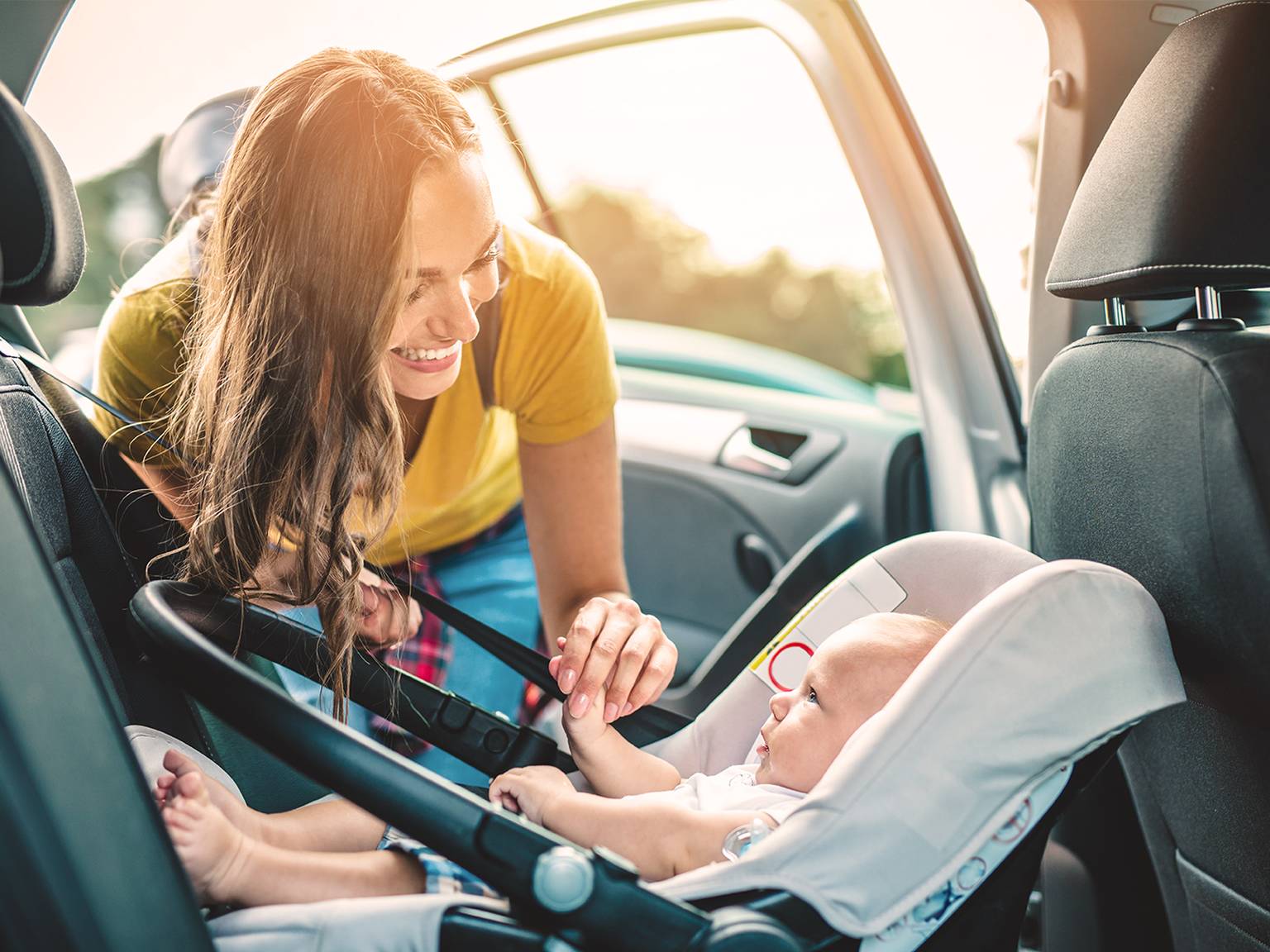 Eine Frau lächelt ein Baby in einem Autositz in einem Fahrzeug an. Die Autotür ist offen, und Sonnenlicht strömt herein.