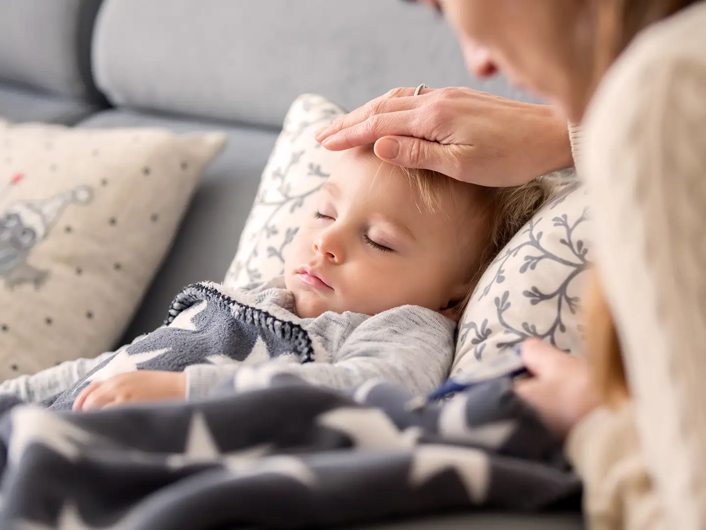 Ein Kind schläft auf einem Sofa unter einer Decke mit Sternenmuster, während ein Erwachsener vorsichtig seine Temperatur mit einem Thermometer misst.
