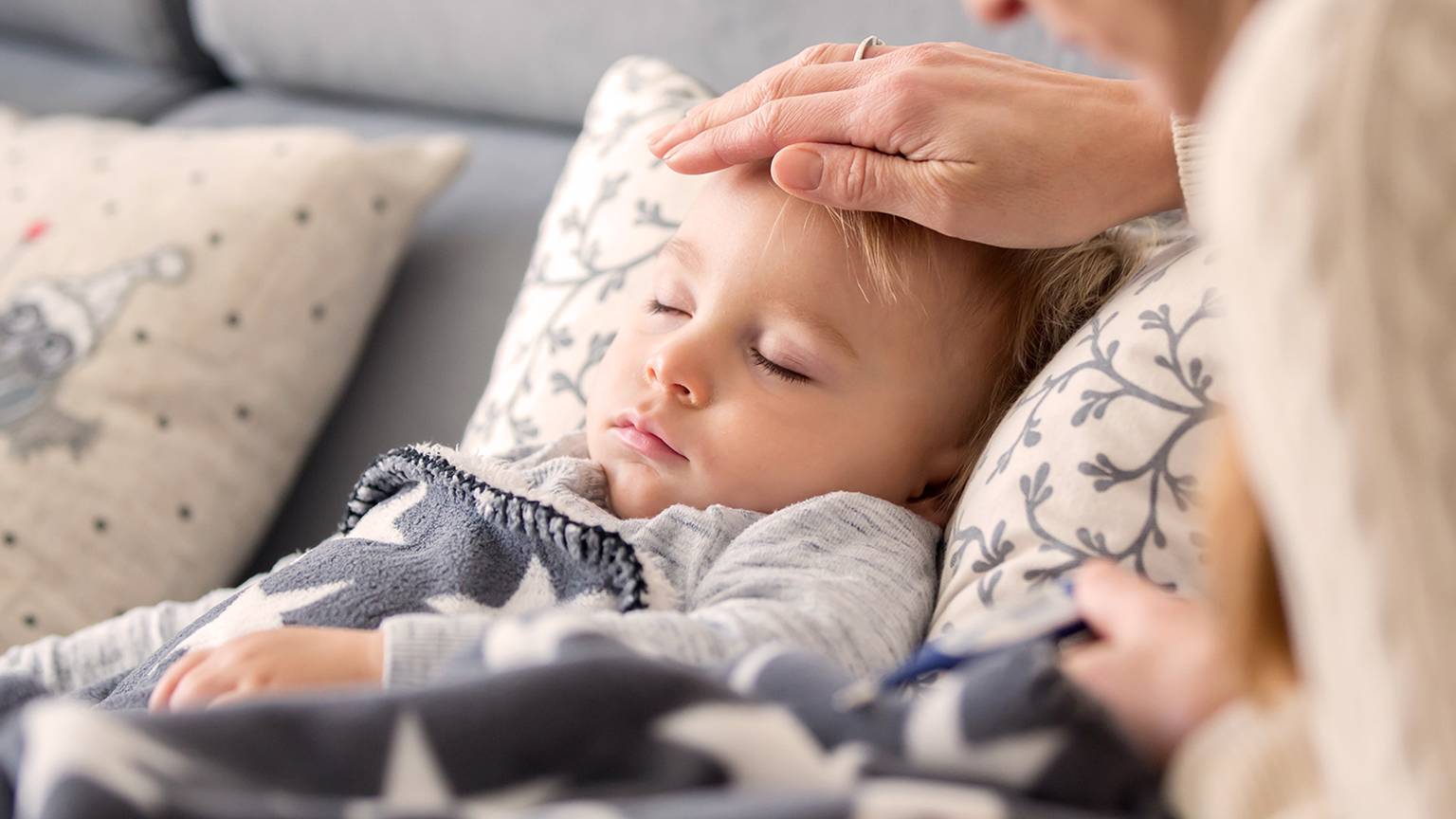 Ein Kind schläft auf einem Sofa unter einer Decke mit Sternenmuster, während ein Erwachsener vorsichtig seine Temperatur mit einem Thermometer misst.