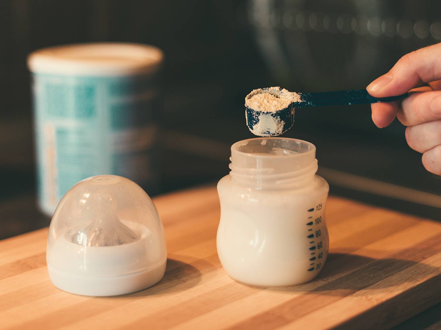 Eine Hand füllt Milchpulver in eine Babyflasche auf einer Holzoberfläche. Daneben stehen ein Behälter und ein Flaschenverschluss.