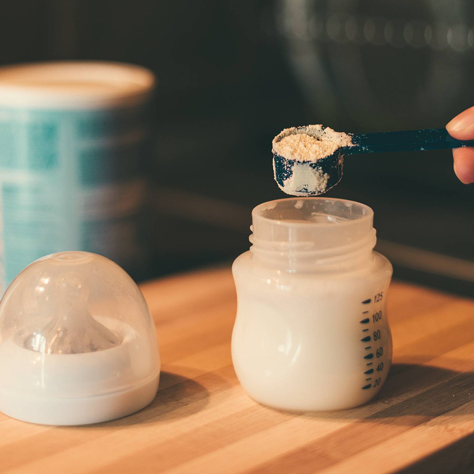 Eine Hand füllt Milchpulver in eine Babyflasche auf einer Holzoberfläche. Daneben stehen ein Behälter und ein Flaschenverschluss.