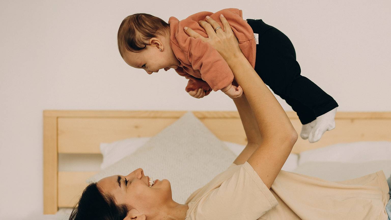 Eine Frau, die auf einem Bett liegt, hebt freudig ein lächelndes Baby über sich. Im Hintergrund sind ein hölzernes Kopfteil und Kissen zu sehen.