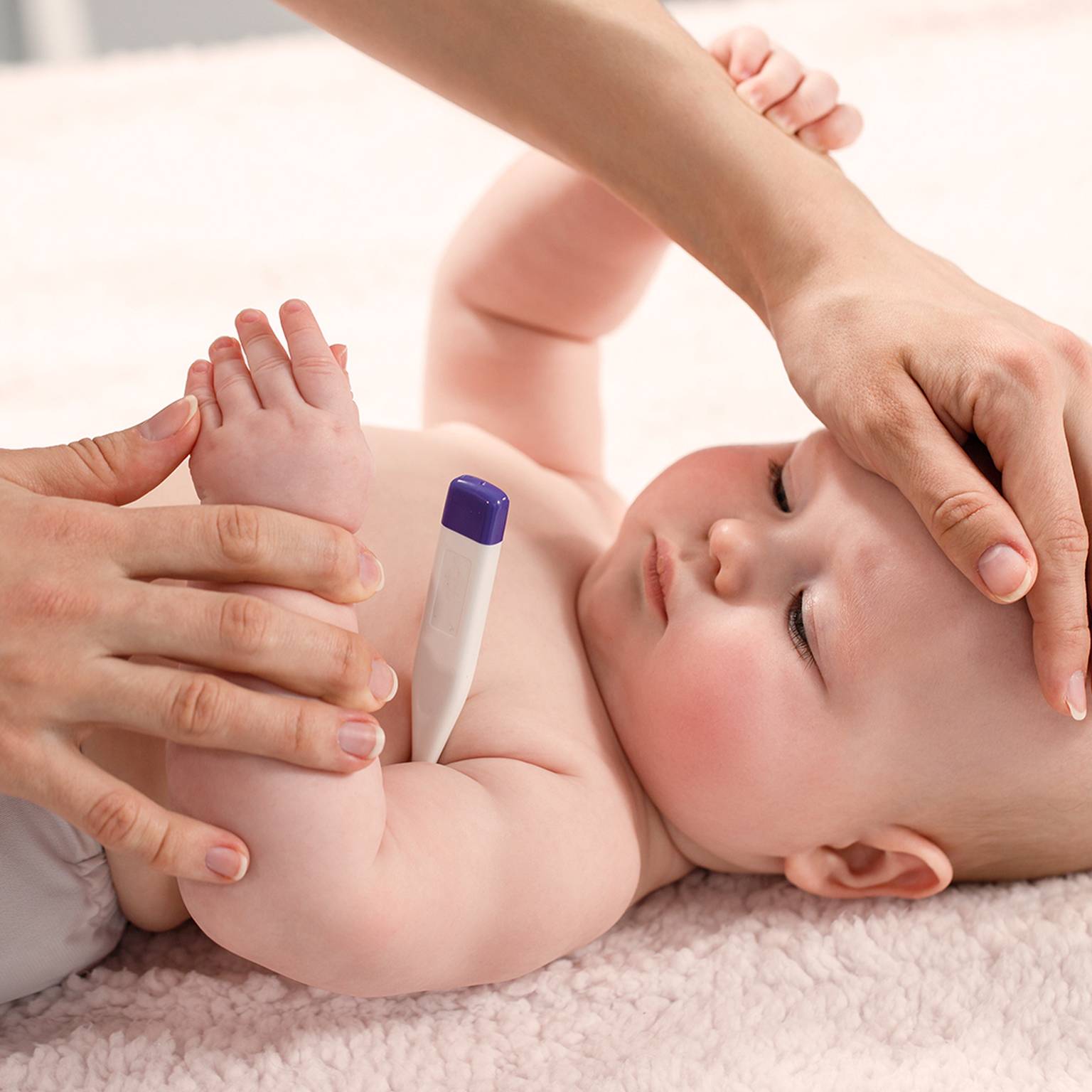 Ein Baby liegt auf einer weichen Oberfläche, während ein Erwachsener seine Temperatur mit einem Thermometer misst und sanft seine Stirn berührt.