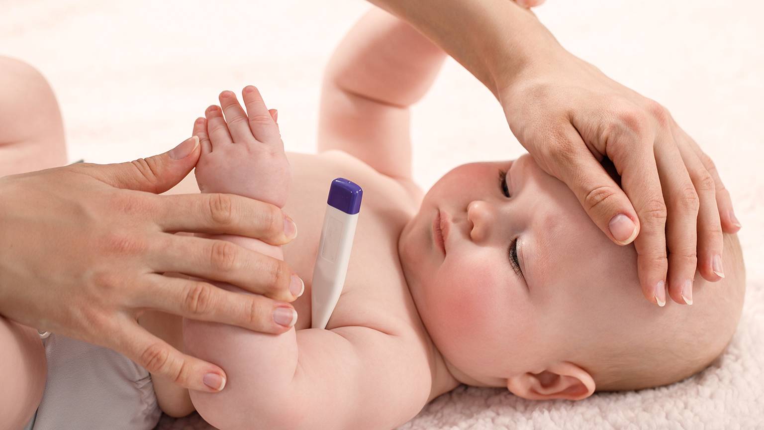 Ein Baby liegt auf einer weichen Oberfläche, während ein Erwachsener seine Temperatur mit einem Thermometer misst und sanft seine Stirn berührt.
