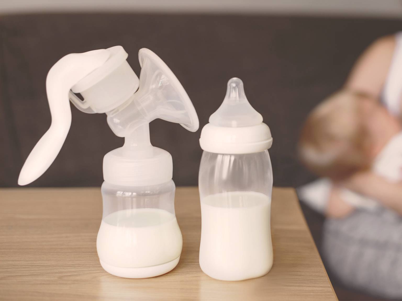 Milchpumpe und mit Milch gefüllte Babyflasche auf einem Tisch, mit einem unscharfen Hintergrund einer Person, die ein Baby hält.