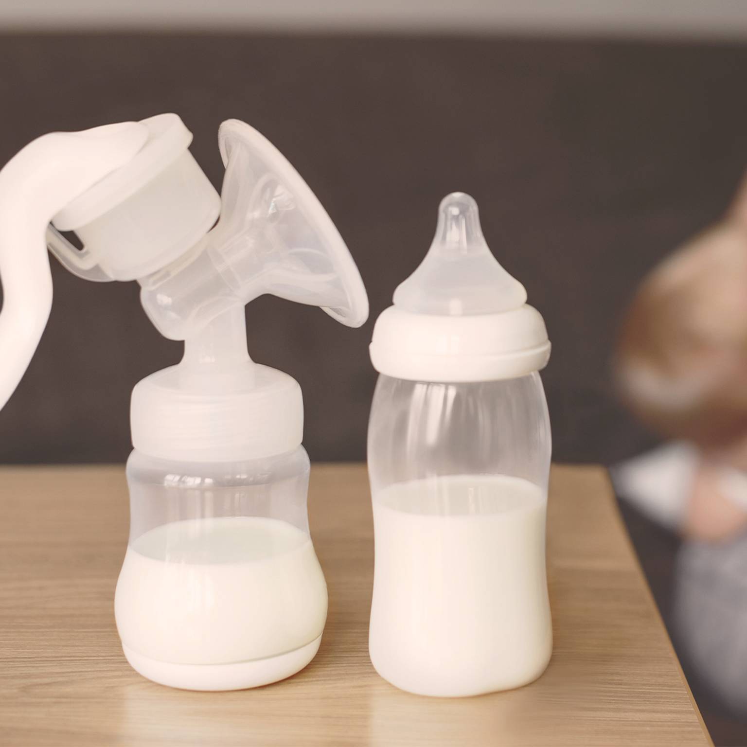 Milchpumpe und mit Milch gefüllte Babyflasche auf einem Tisch, mit einem unscharfen Hintergrund einer Person, die ein Baby hält.
