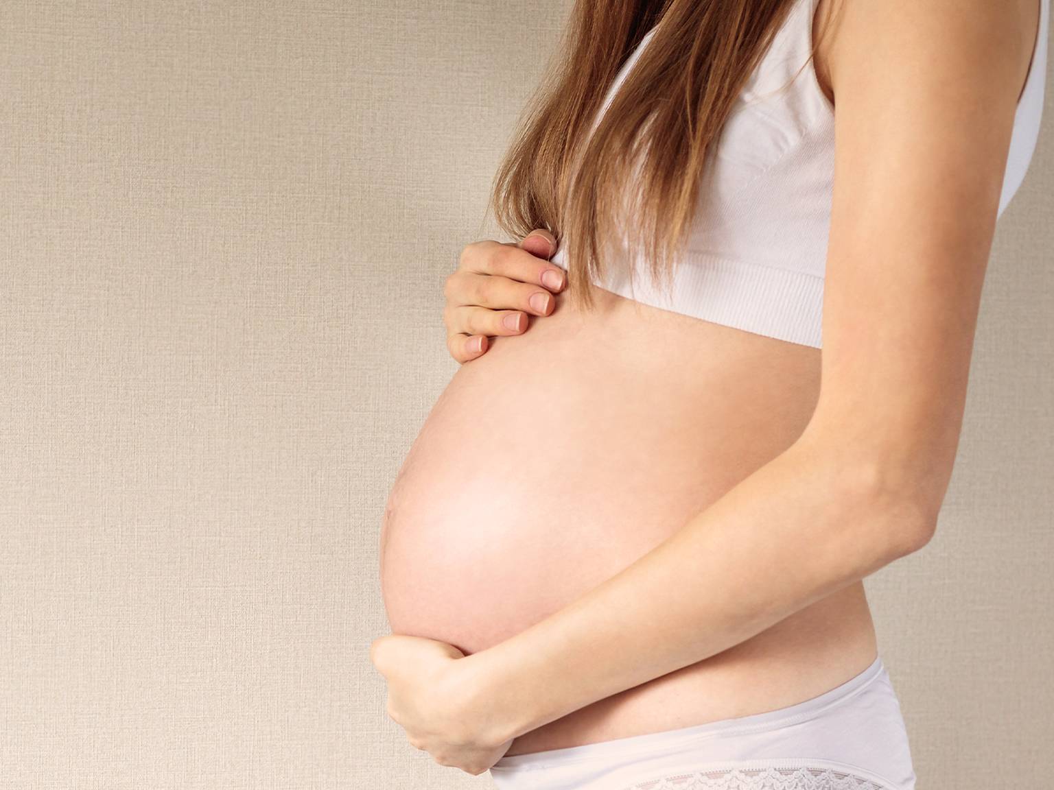 Schwangere Frau in weißem Oberteil und Unterwäsche, die vor neutralem Hintergrund sanft ihren Bauch mit beiden Händen hält.