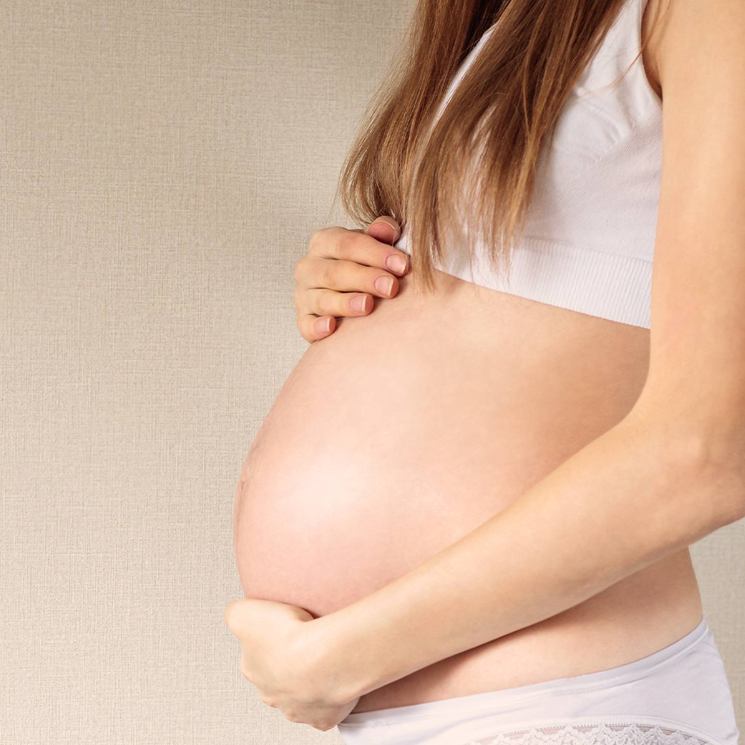 Schwangere Frau in weißem Oberteil und Unterwäsche, die vor neutralem Hintergrund sanft ihren Bauch mit beiden Händen hält.