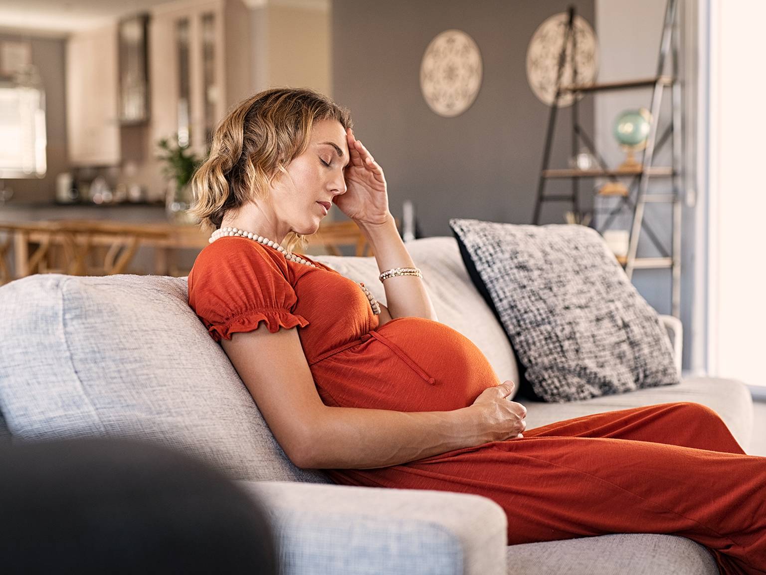 Schwangere Frau in einem roten Kleid sitzt auf einem Sofa, hält mit einer Hand ihren Kopf und mit der anderen ihren Bauch und sieht müde aus.