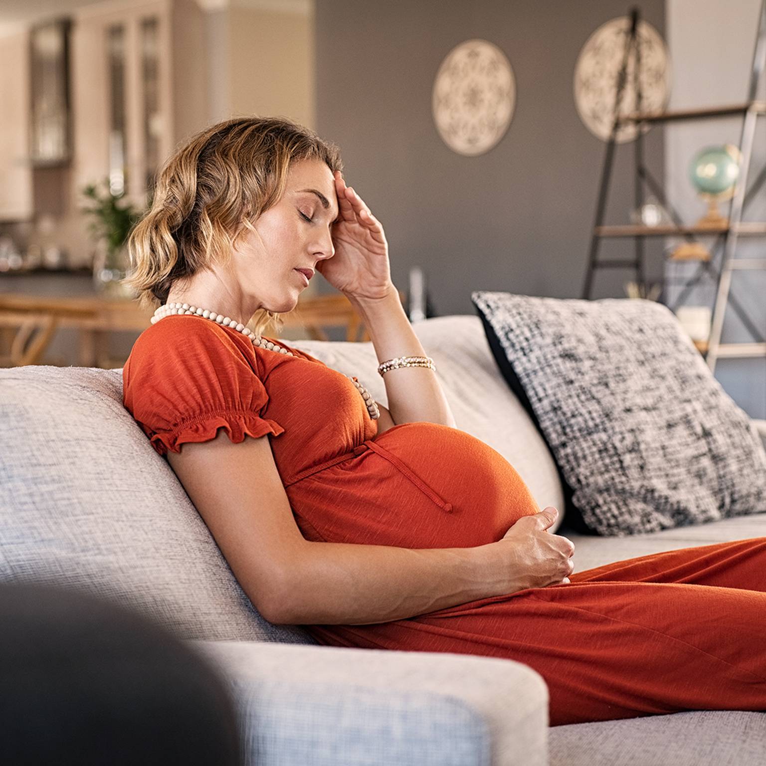 Schwangere Frau in einem roten Kleid sitzt auf einem Sofa, hält mit einer Hand ihren Kopf und mit der anderen ihren Bauch und sieht müde aus.