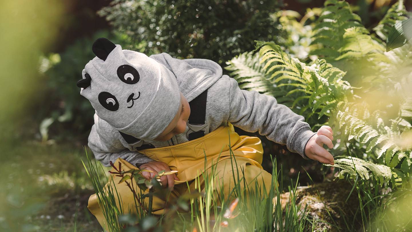Kind mit Pandamütze und gelbem Overall erkundet Pflanzen in einem Garten, umgeben von Grün und Sonnenlicht.
