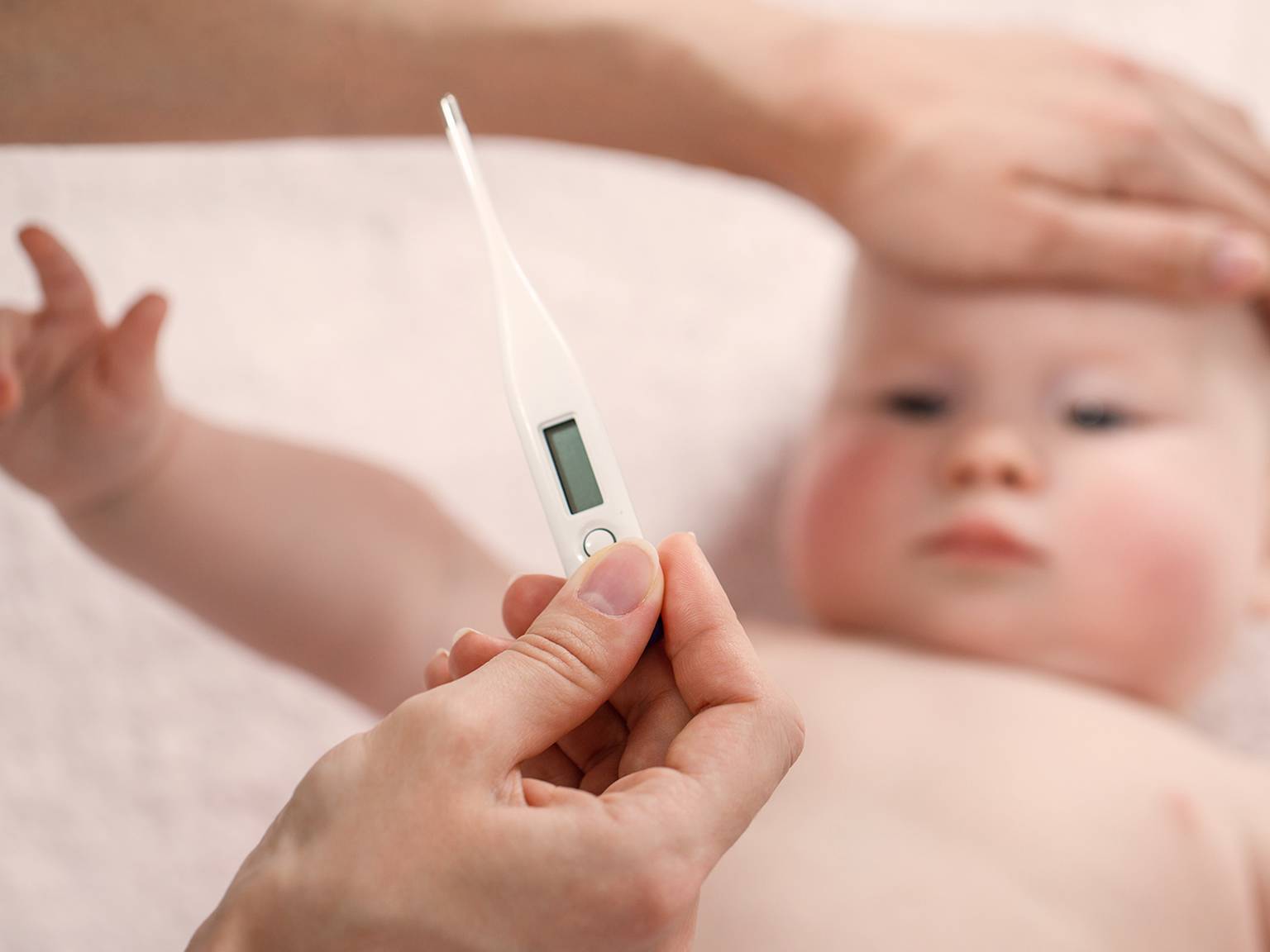 Eine Person hält ein digitales Thermometer an die Stirn eines Babys, während eine andere Hand sanft den Kopf des Babys berührt, um zu prüfen, ob es Fieber hat.