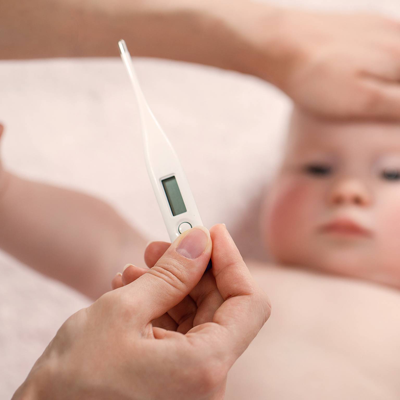 Eine Person hält ein digitales Thermometer an die Stirn eines Babys, während eine andere Hand sanft den Kopf des Babys berührt, um zu prüfen, ob es Fieber hat.