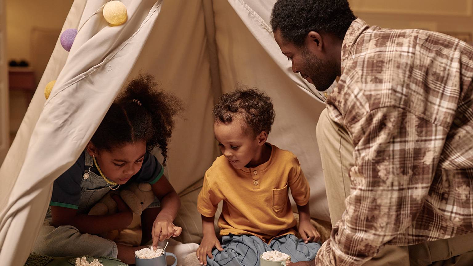 Ein Vater und zwei Kinder sitzen in einem gemütlichen Zelt und trinken heiße Schokolade mit Marshmallows. Das Zelt ist mit Lichterketten dekoriert.