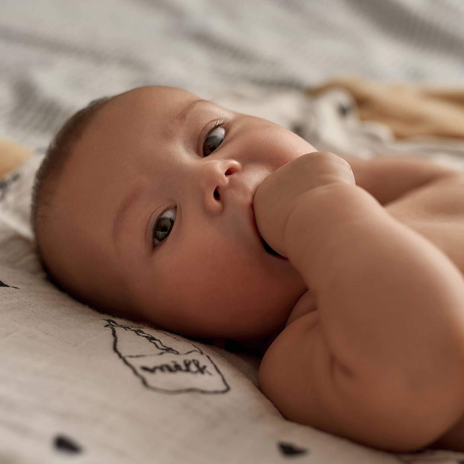 Baby liegt auf einer gemusterten Decke, schaut nach oben und hat eine Hand im Mund.