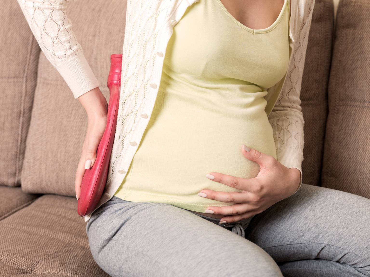 Schwangere Frau auf einer Couch, die mit einer Hand ihren Bauch hält und mit der anderen eine rote Wärmflasche auf ihrem Rücken hält.