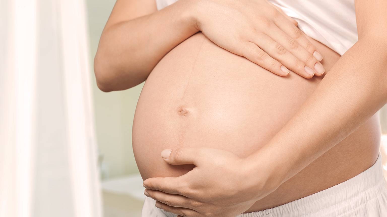 Eine schwangere Frau in weißem Hemd und Shorts streichelt sanft mit beiden Händen ihren nackten Bauch in einem schwach beleuchteten Raum.