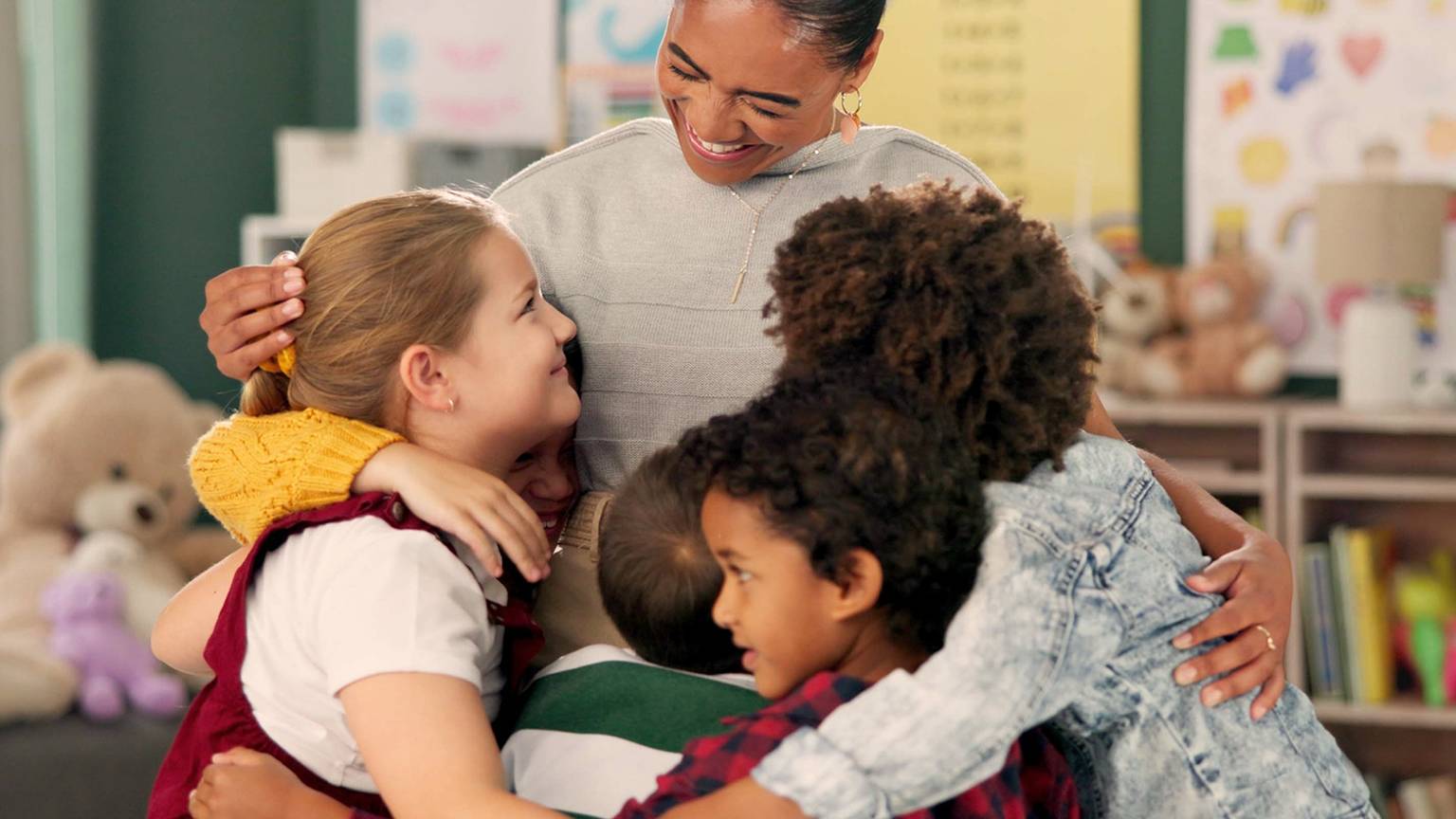 Eine Lehrerin umarmt herzlich eine bunt gemischte Gruppe von Kindern in einem Klassenzimmer, umgeben von bunten Postern und Stofftieren.