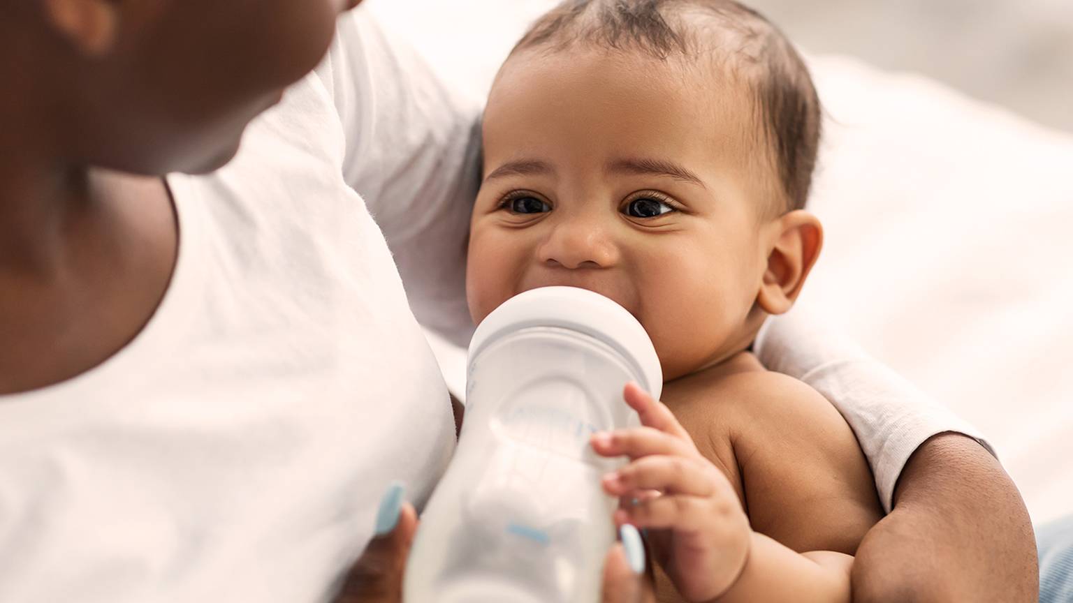 Ein Baby trinkt aus einer Flasche, während es von einem Erwachsenen gehalten wird. Das Baby sieht zufrieden aus und liegt in den Armen des Erwachsenen.
