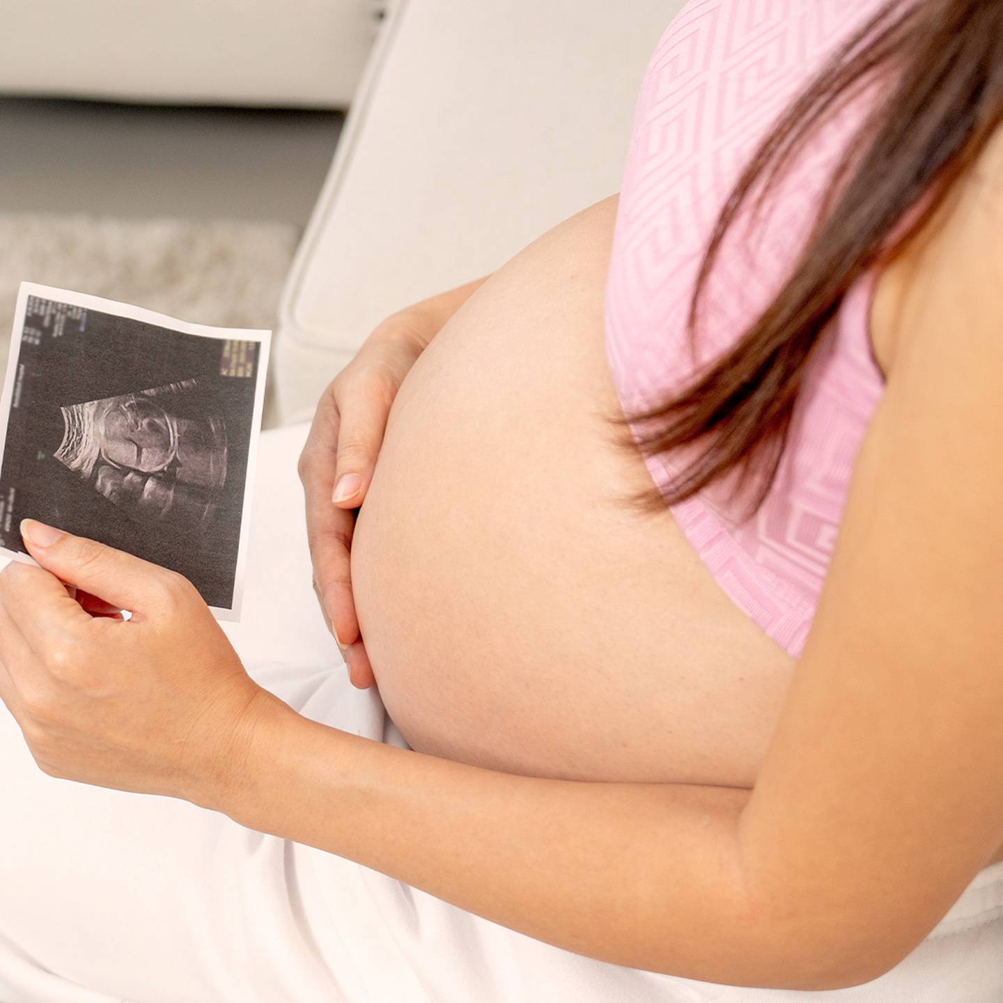 Schwangere Frau in einem rosa Oberteil hält ein Ultraschallbild in der Hand, sitzt auf einer Couch und zeigt ihren Babybauch.