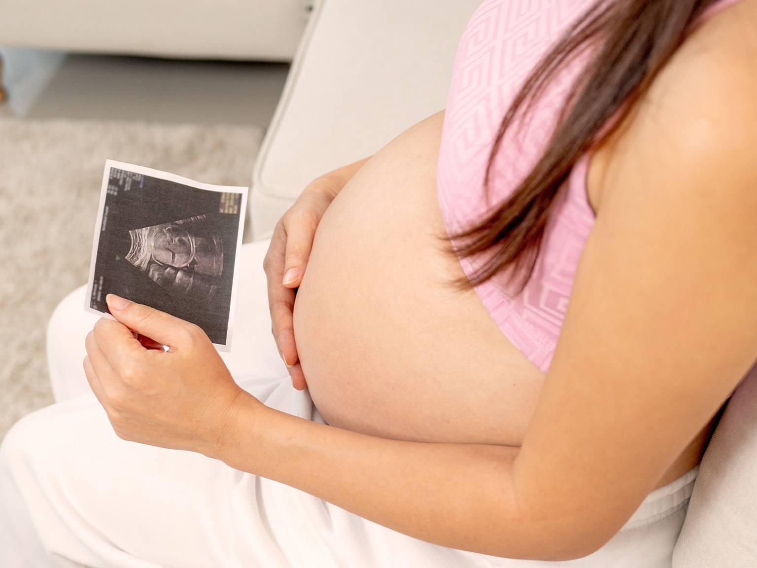 Schwangere Frau in einem rosa Oberteil hält ein Ultraschallbild in der Hand, sitzt auf einer Couch und zeigt ihren Babybauch.