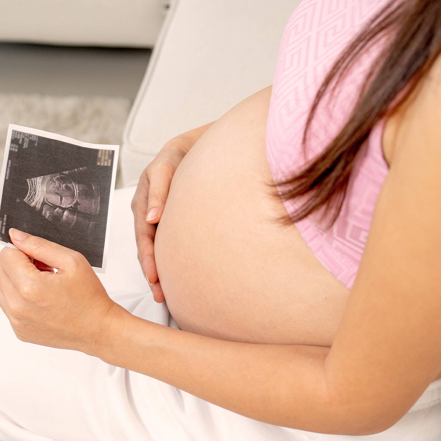 Schwangere Frau in einem rosa Oberteil hält ein Ultraschallbild in der Hand, sitzt auf einer Couch und zeigt ihren Babybauch.