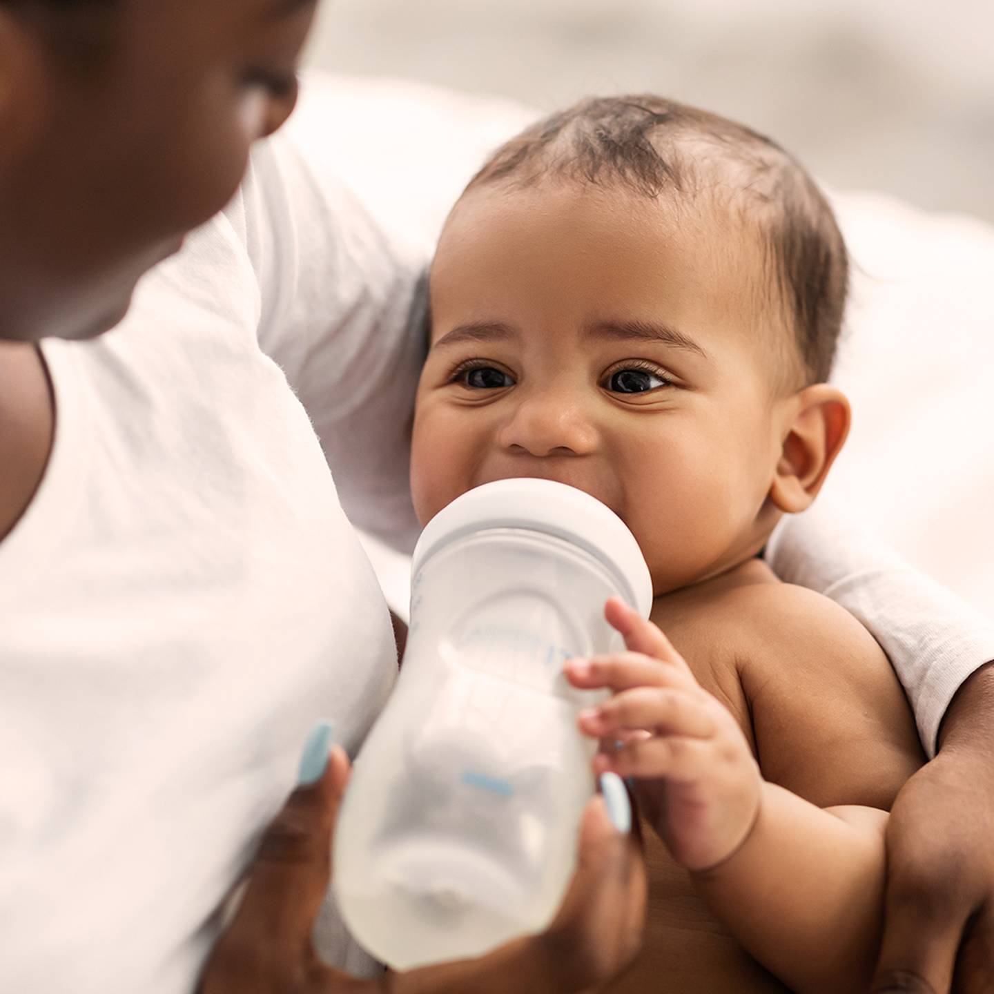 Ein Baby trinkt aus einer Flasche, während es von einem Erwachsenen gehalten wird. Beide sitzen auf einem Bett. Das Baby sieht zufrieden und entspannt aus.