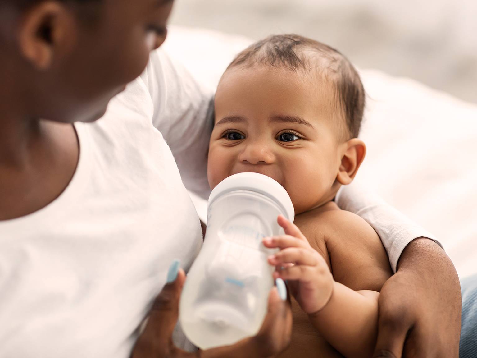Ein Baby trinkt aus einer Flasche, während es von einem Erwachsenen gehalten wird. Beide sitzen auf einem Bett. Das Baby sieht zufrieden und entspannt aus.