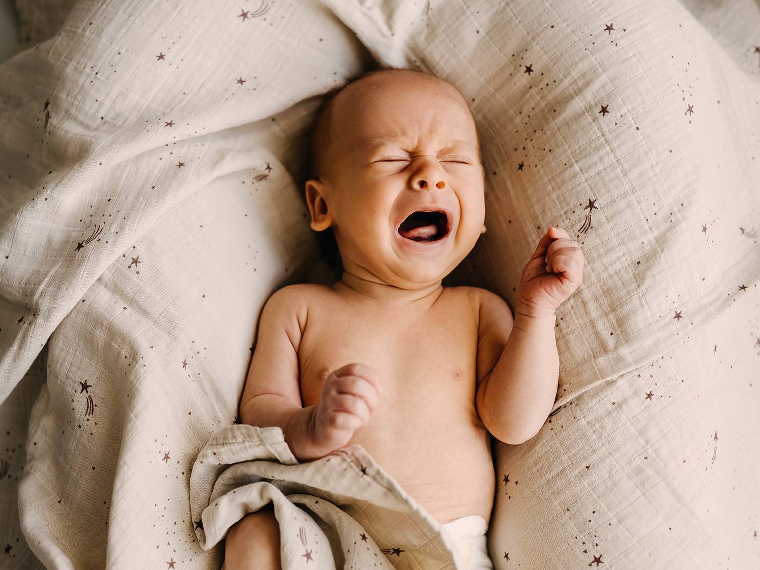 Ein weinendes Baby liegt mit geschlossenen Augen und geballten Fäusten auf einer weichen Decke mit Sternenmuster.