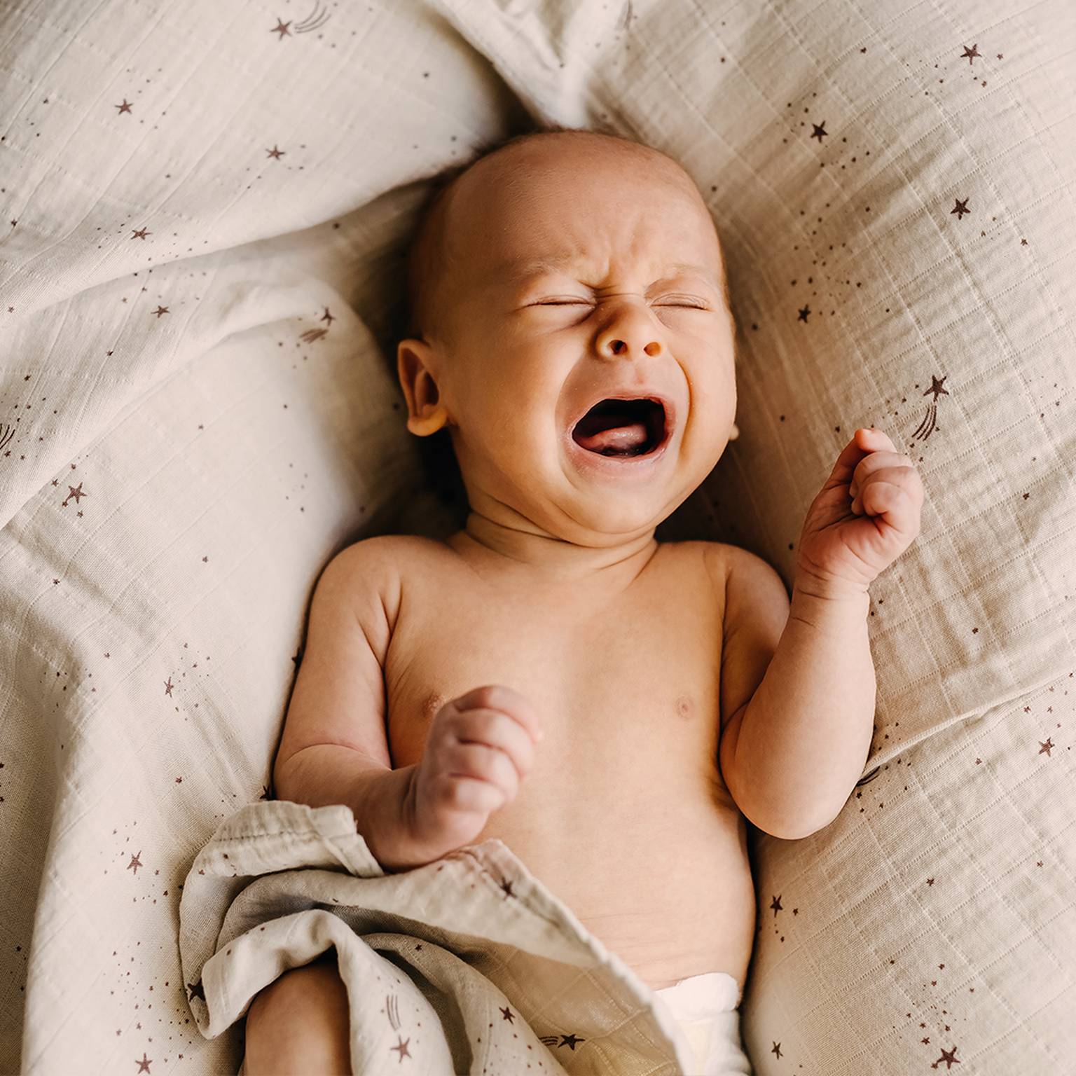 Ein weinendes Baby liegt mit geschlossenen Augen und geballten Fäusten auf einer weichen Decke mit Sternenmuster.