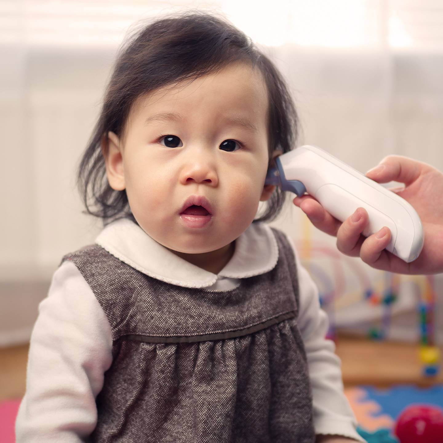 Ein Baby in einem grauen Kleid sitzt auf einer bunten Matte, während ein Erwachsener mit einem Ohrthermometer die Temperatur des Babys misst.