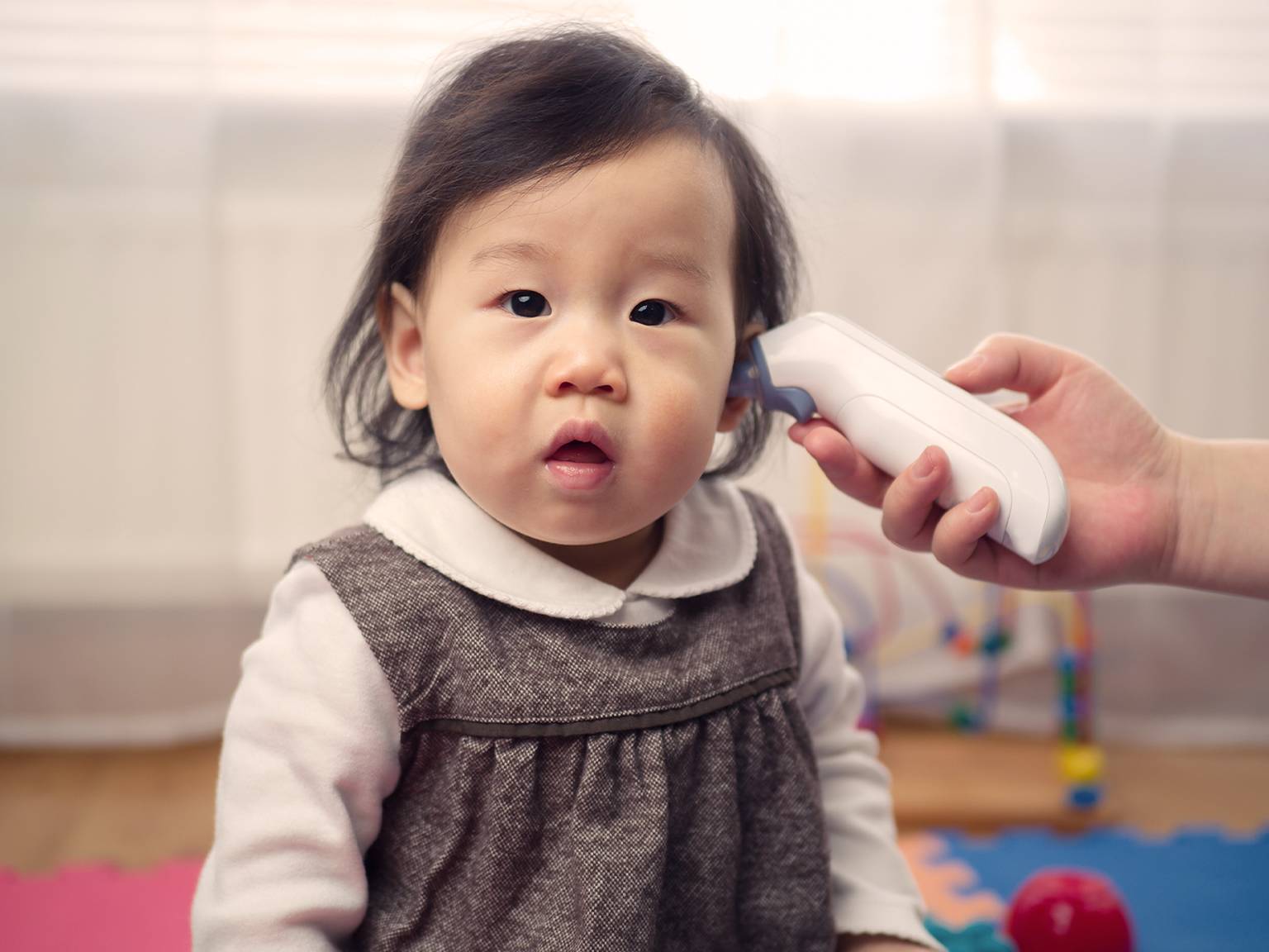 Ein Baby in einem grauen Kleid sitzt auf einer bunten Matte, während ein Erwachsener mit einem Ohrthermometer die Temperatur des Babys misst.