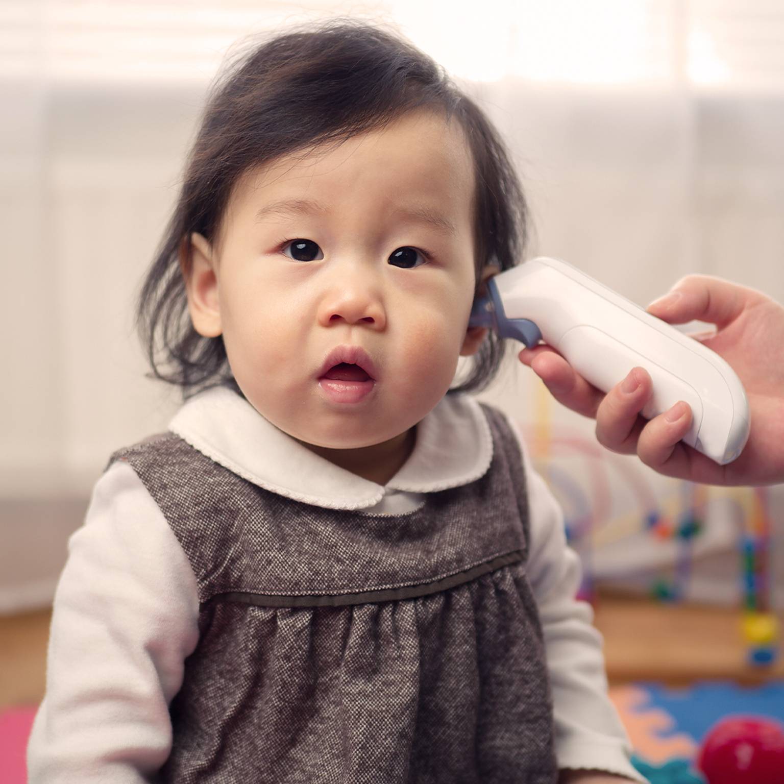 Ein Baby in einem grauen Kleid sitzt auf einer bunten Matte, während ein Erwachsener mit einem Ohrthermometer die Temperatur des Babys misst.