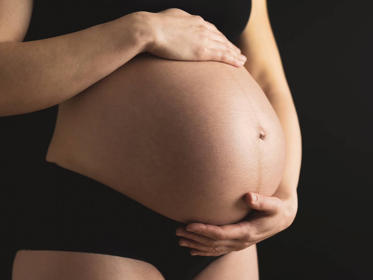 Schwangere Person in schwarzer Unterwäsche wiegt ihren Bauch mit beiden Händen vor einem dunklen Hintergrund.