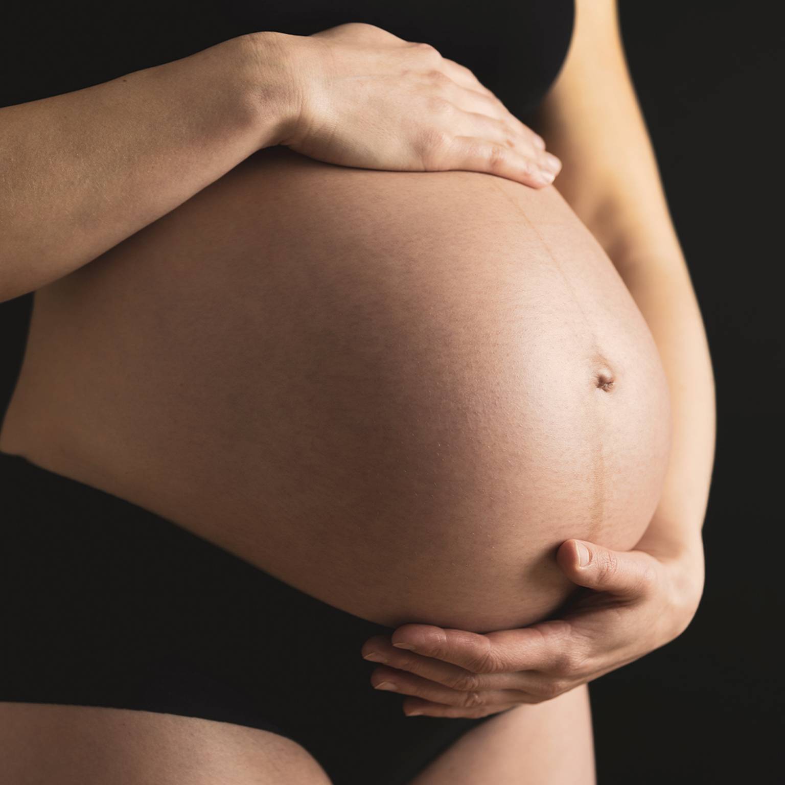 Schwangere Person in schwarzer Unterwäsche wiegt ihren Bauch mit beiden Händen vor einem dunklen Hintergrund.