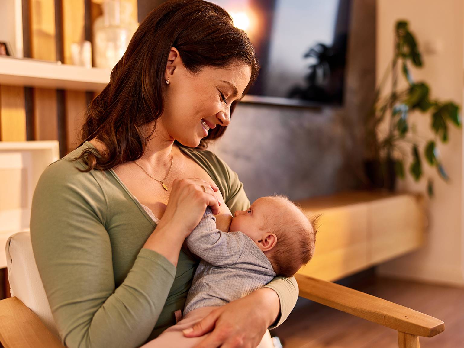 Eine Frau in einem grünen Oberteil lächelt, während sie in einem gemütlichen, warm beleuchteten Raum mit Pflanzen und Holzmöbeln ein Baby hält und stillt.