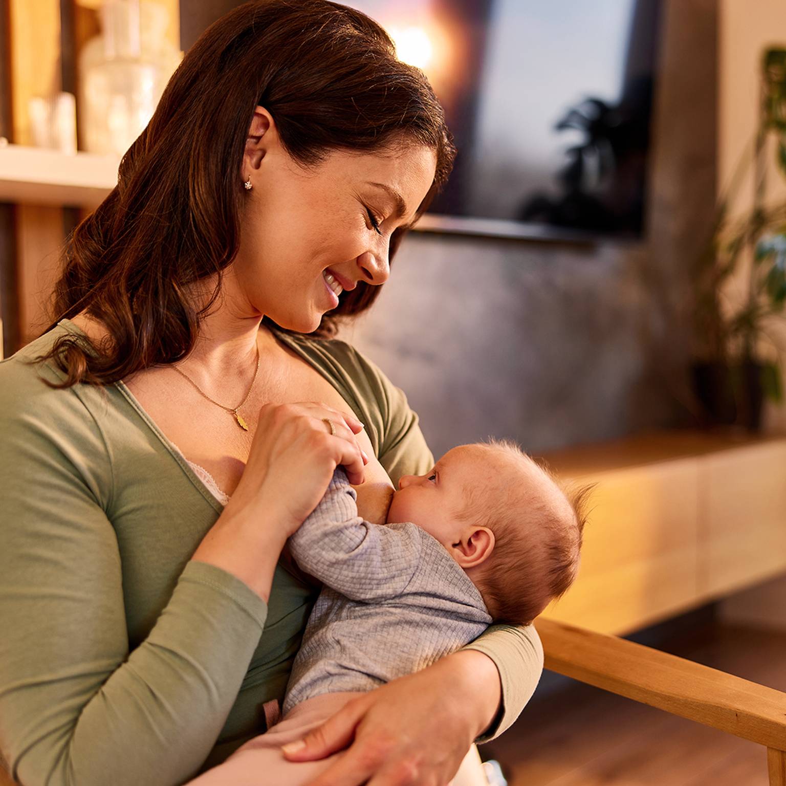 Eine Frau in einem grünen Oberteil lächelt, während sie in einem gemütlichen, warm beleuchteten Raum mit Pflanzen und Holzmöbeln ein Baby hält und stillt.