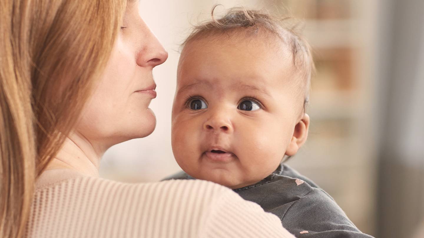 Frau hält ein Baby im Arm, beide blicken nach vorne. Das Baby schaut neugierig geradeaus, während die Frau zur Seite blickt.