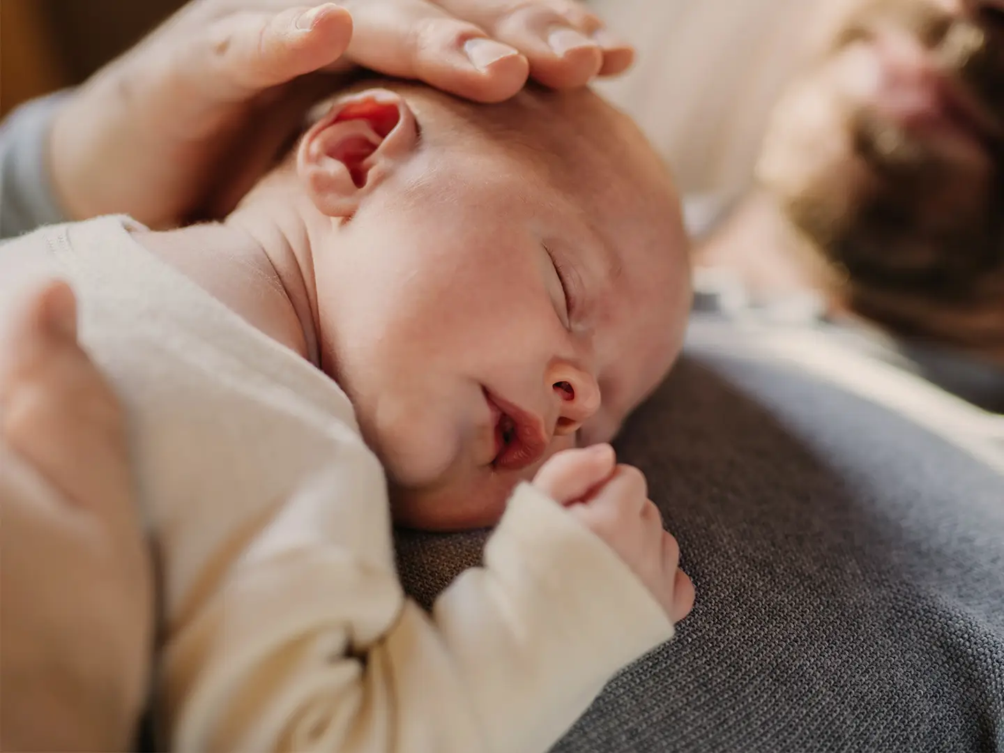 Ein schlafendes Neugeborenes ruht auf der Brust eines Erwachsenen, während eine sanfte Hand den Kopf des Babys wiegt und so einen ruhigen und zärtlichen Moment schafft.