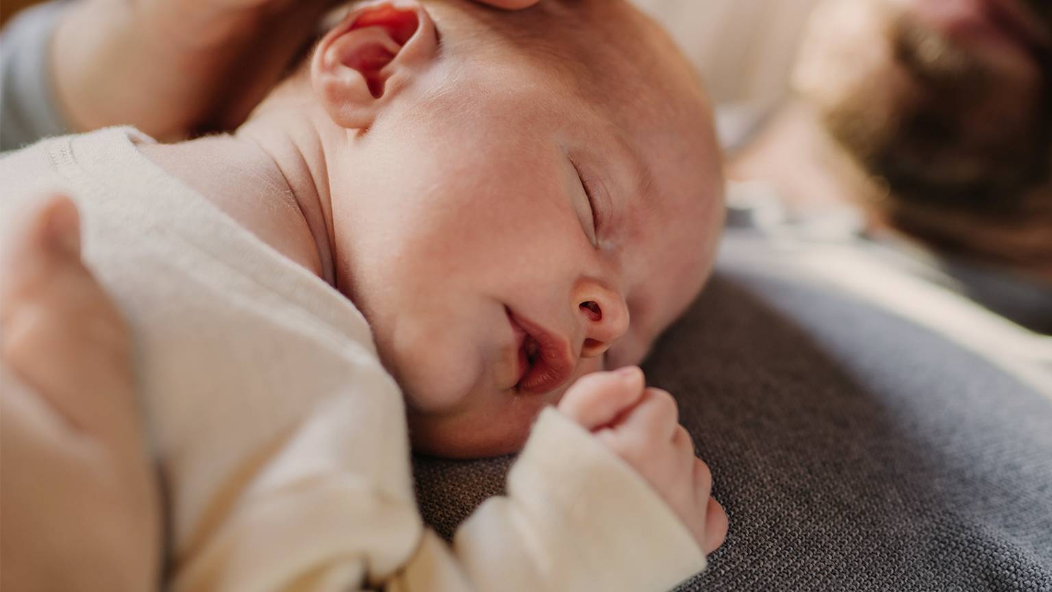 Ein schlafendes Neugeborenes ruht auf der Brust eines Erwachsenen, während eine sanfte Hand den Kopf des Babys wiegt und so einen ruhigen und zärtlichen Moment schafft.