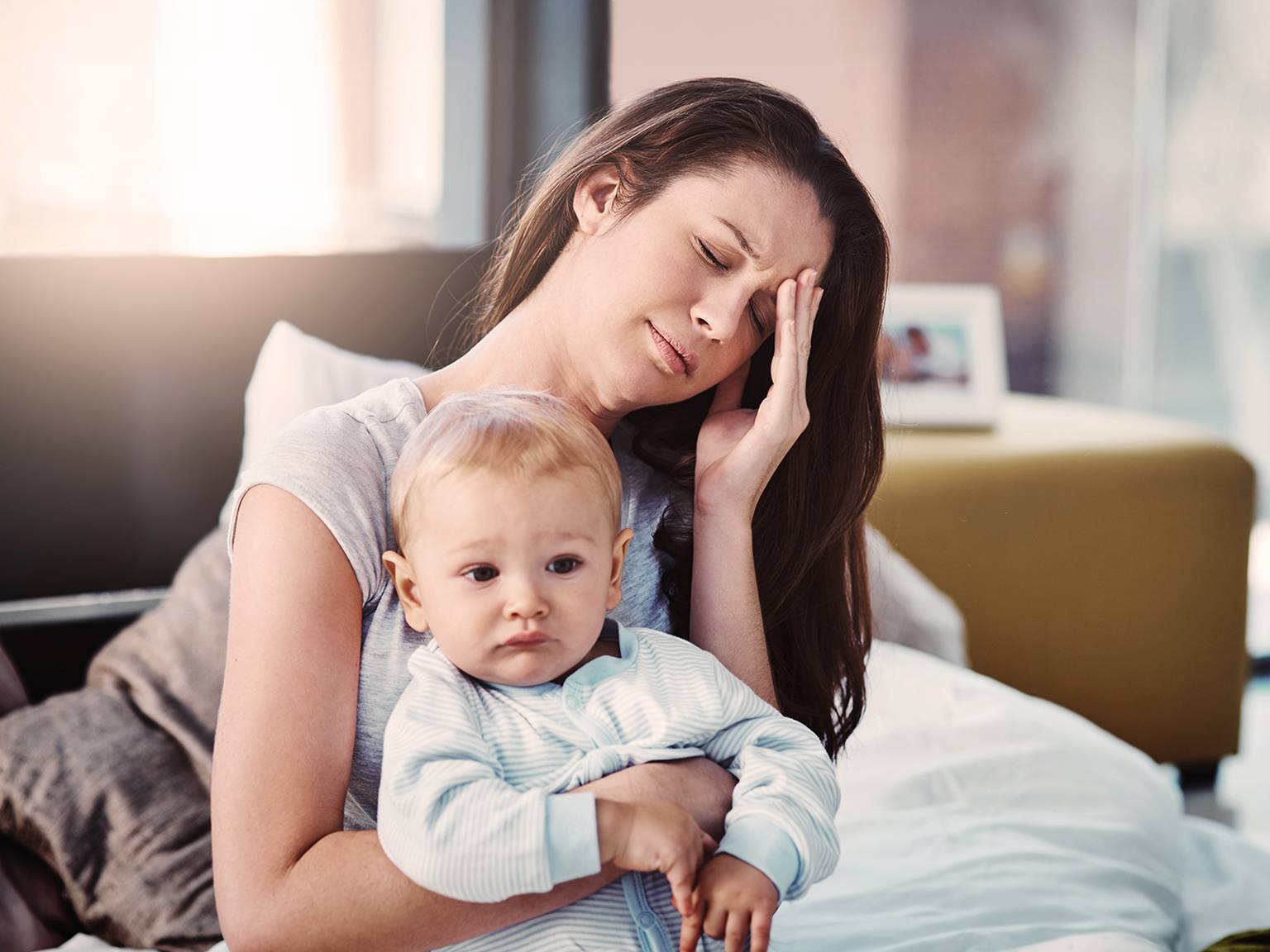 Eine müde Frau, die auf einem Bett sitzt, ein Baby im Arm hält, die Hand auf der Stirn hat und gestresst aussieht.